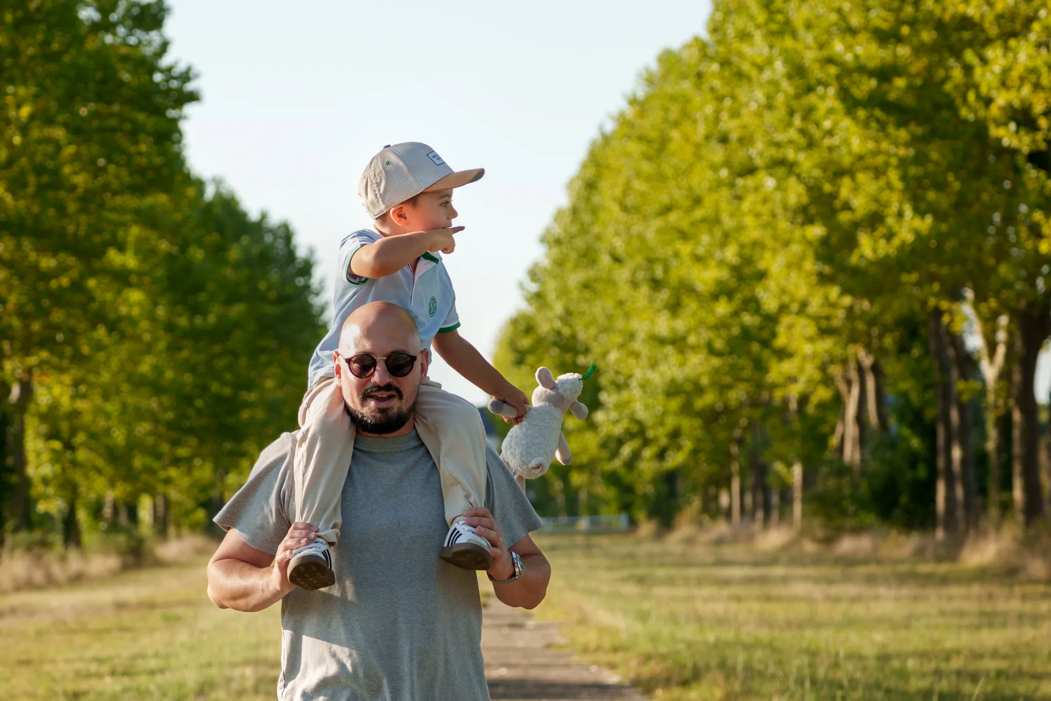 Petit garçon et son doudou, sur les épaules du papa, sur un chemin bordé d'arbres.
