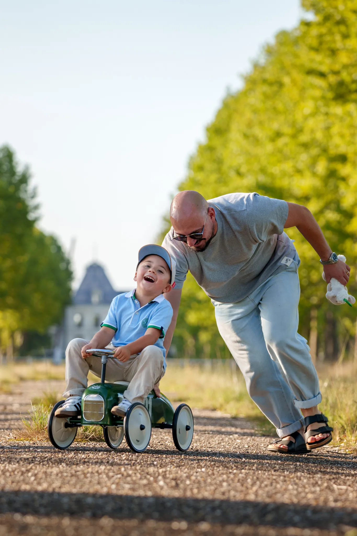Père poussant son fils heureux sur une voiture à pédales verte, sur un chemin arboré.